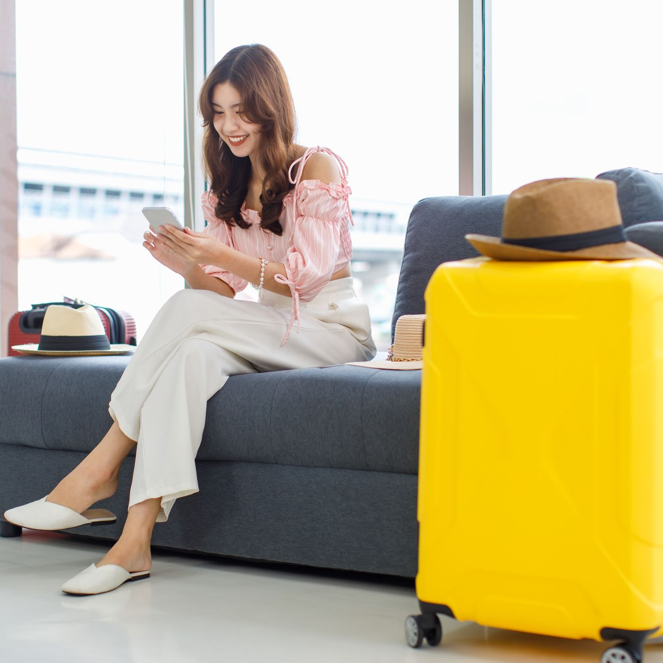 Femme assise sur un canapé, utilisant un téléphone ; valise jaune, chapeau à proximité.