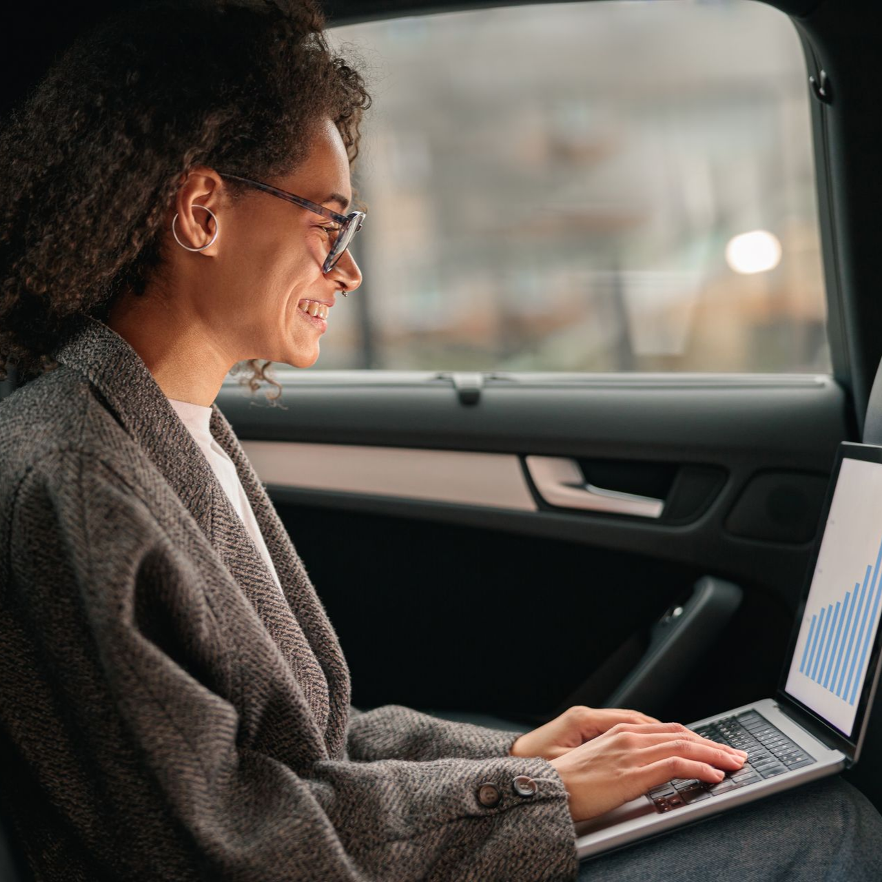 Femme en voiture, travaillant sur un ordinateur portable, souriante. Graphique à l'écran, veste grise.