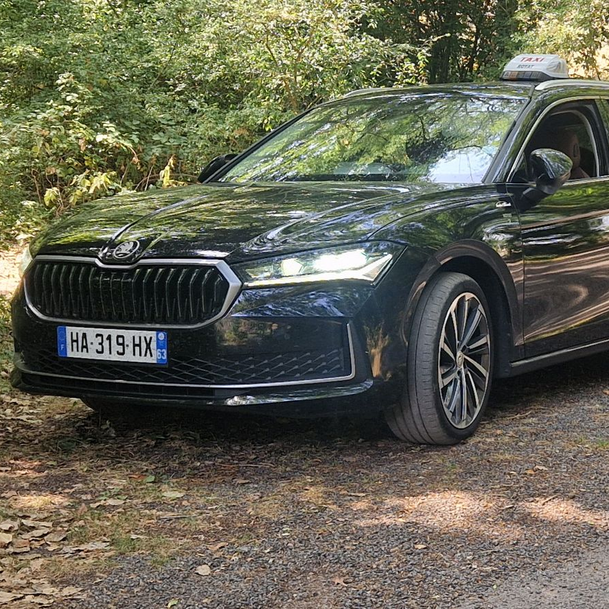 Un taxi Skoda noir immatriculé en France, stationné sur un chemin de terre devant des arbres.