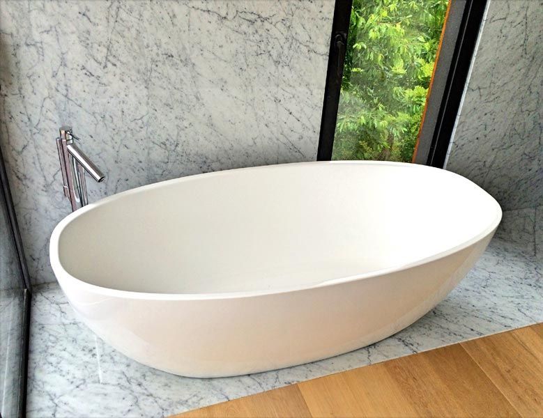 Baignoire ovale blanche posée sur du marbre, près d'une fenêtre donnant sur la verdure. Robinet chromé à gauche.