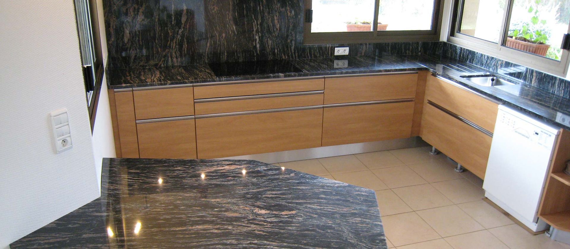 Cuisine avec placards en bois, plans de travail noirs et carrelage clair au sol.