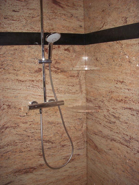 Douche avec murs en granit beige et rouge, pommeau et robinetterie chromés, et étagères intégrées.