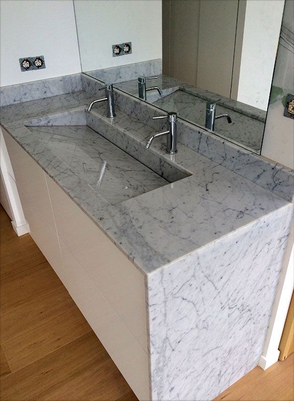 Meuble-lavabo moderne avec vasque en marbre intégrée et trois robinets. Armoires blanches, miroir et parquet.