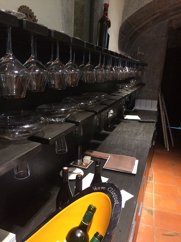 Bar avec verres suspendus et bouteilles sur étagères. Intérieur en bois sombre et pierre.