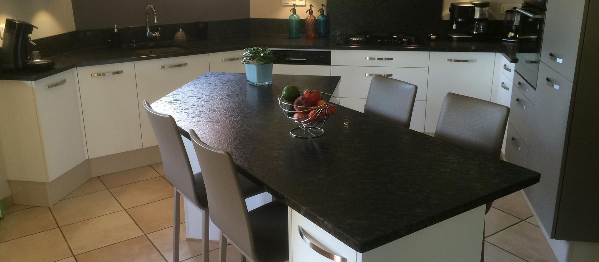 Cuisine avec un plan de travail noir, des placards blancs et une table avec des fruits et des chaises.