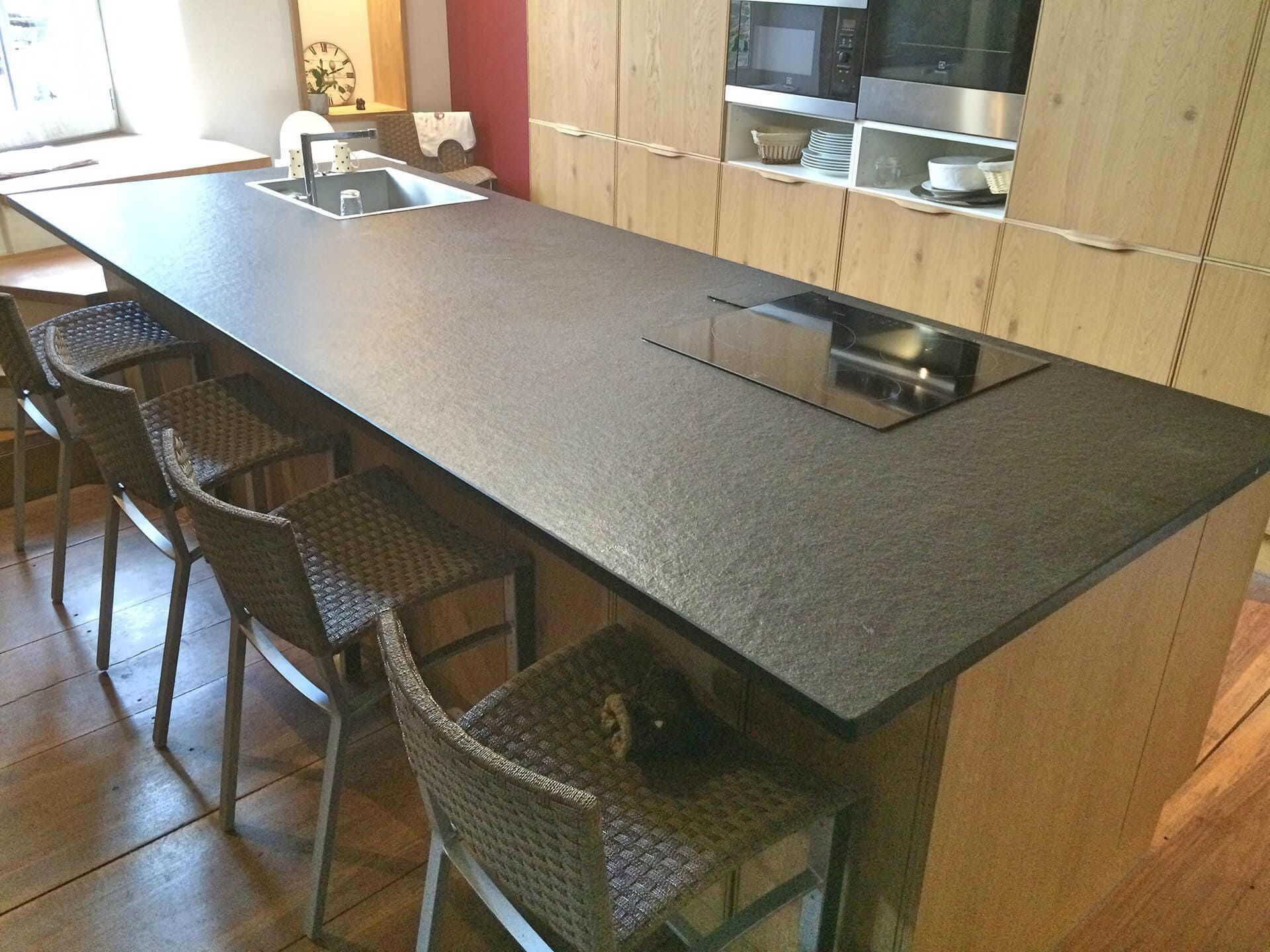 Îlot de cuisine avec plan de travail foncé, plaque de cuisson et évier intégrés, entouré de tabourets de bar en osier. Placards et sol en bois.