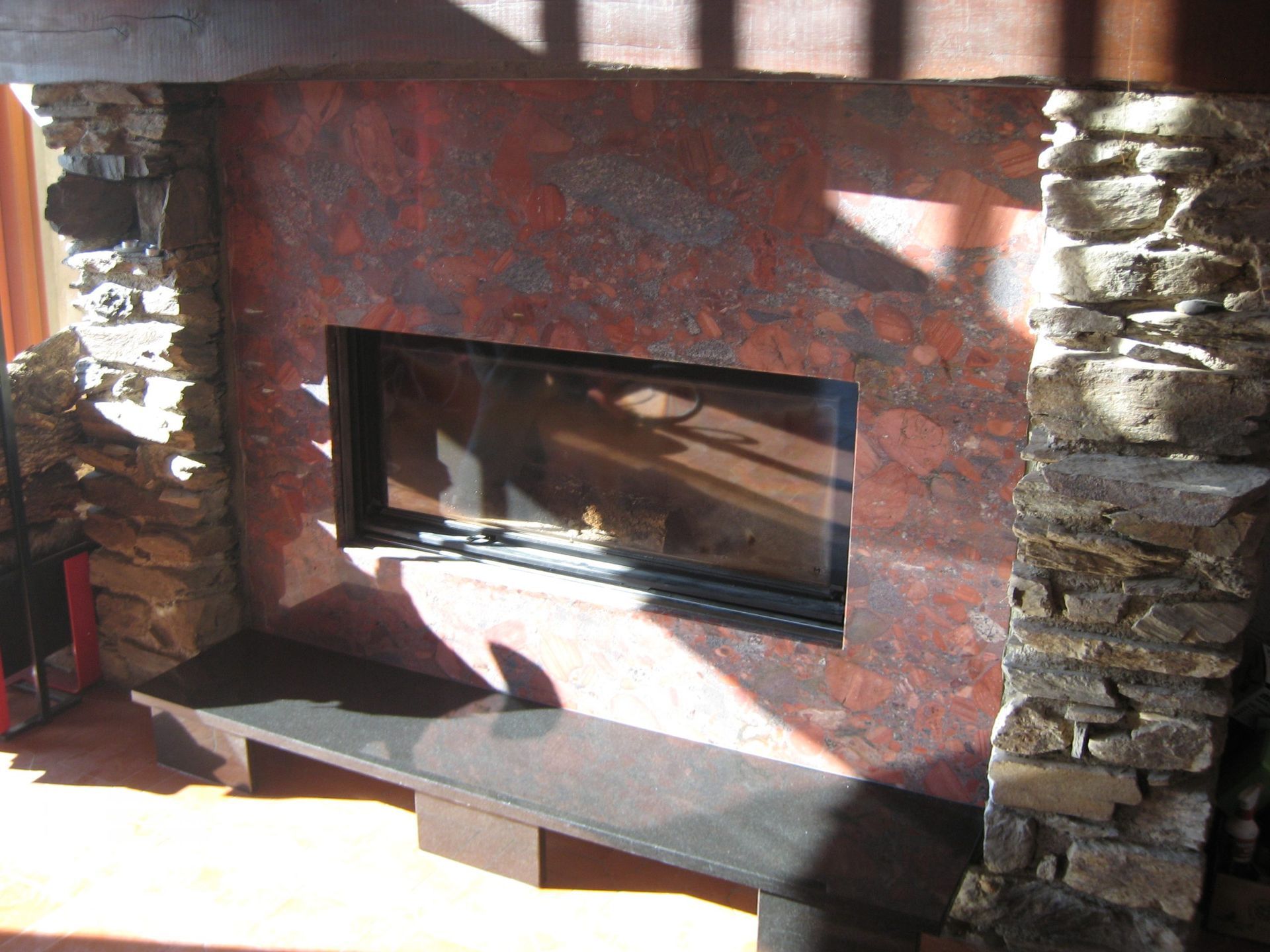 Cheminée avec colonnes en pierre et mur texturé rouge/brun. Banc noir.