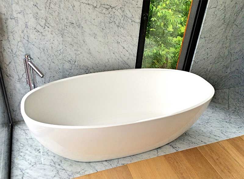 Baignoire ovale blanche sur marbre, près d'une fenêtre avec vue sur la verdure. Robinetterie chromée murale.