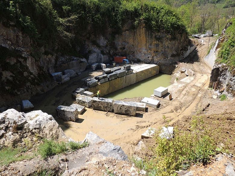 Carrière avec de gros blocs de pierre, de l'eau boueuse et des machines. Un ouvrier se tient à proximité, dans un cadre naturel.