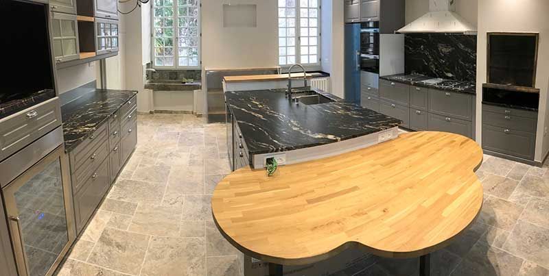 Cuisine avec îlot central, comptoirs en granit noir, table en bois, armoires grises, fenêtre.