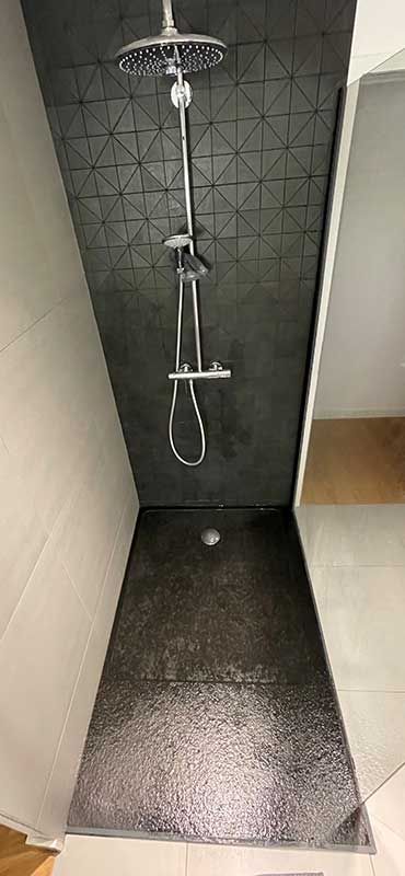 Douche avec murs carrelés noirs, pommeau de douche et sol avec bonde.