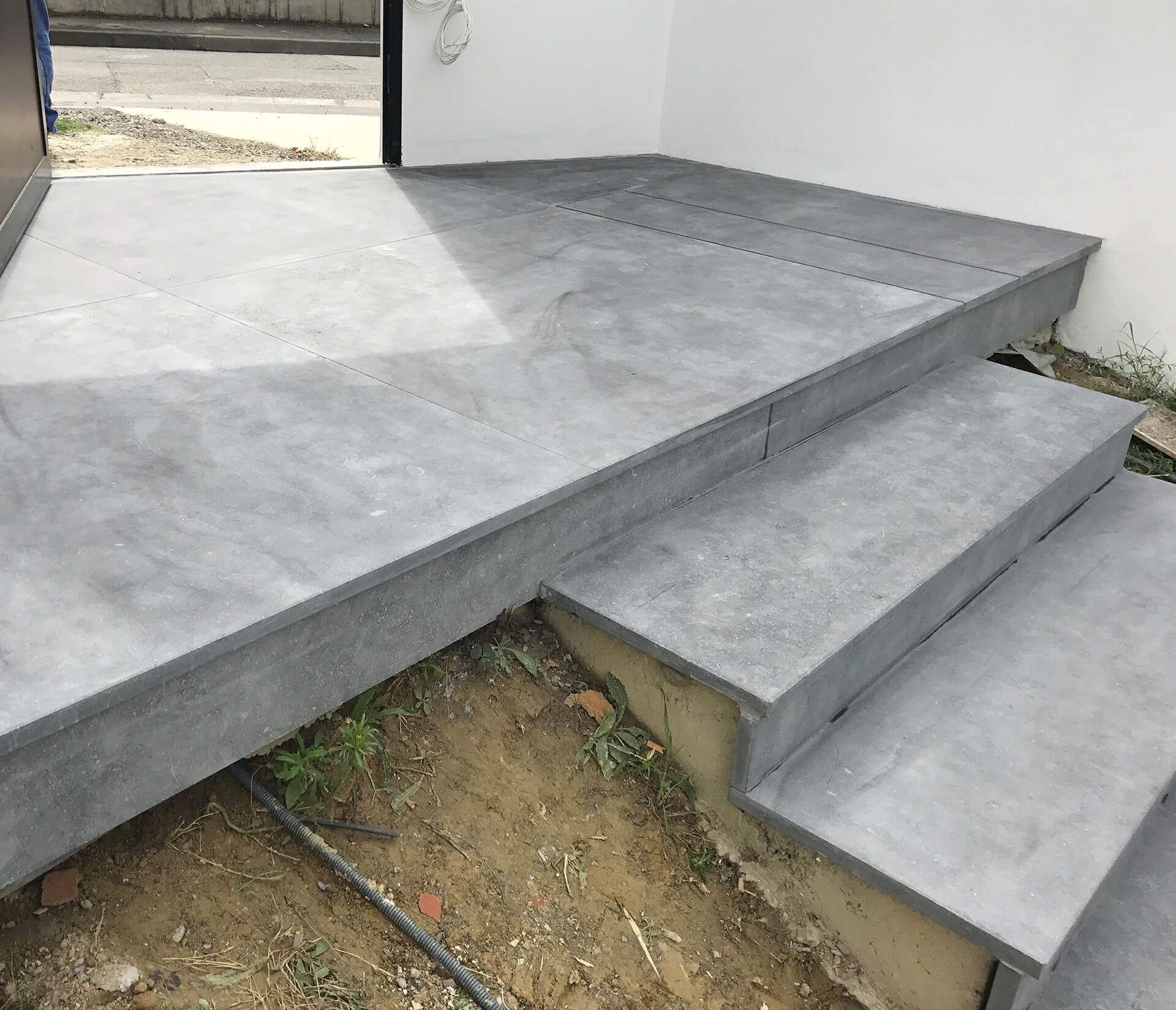 Marches et palier en pierre grise devant un bâtiment. La lumière du soleil projette des ombres sur la surface.