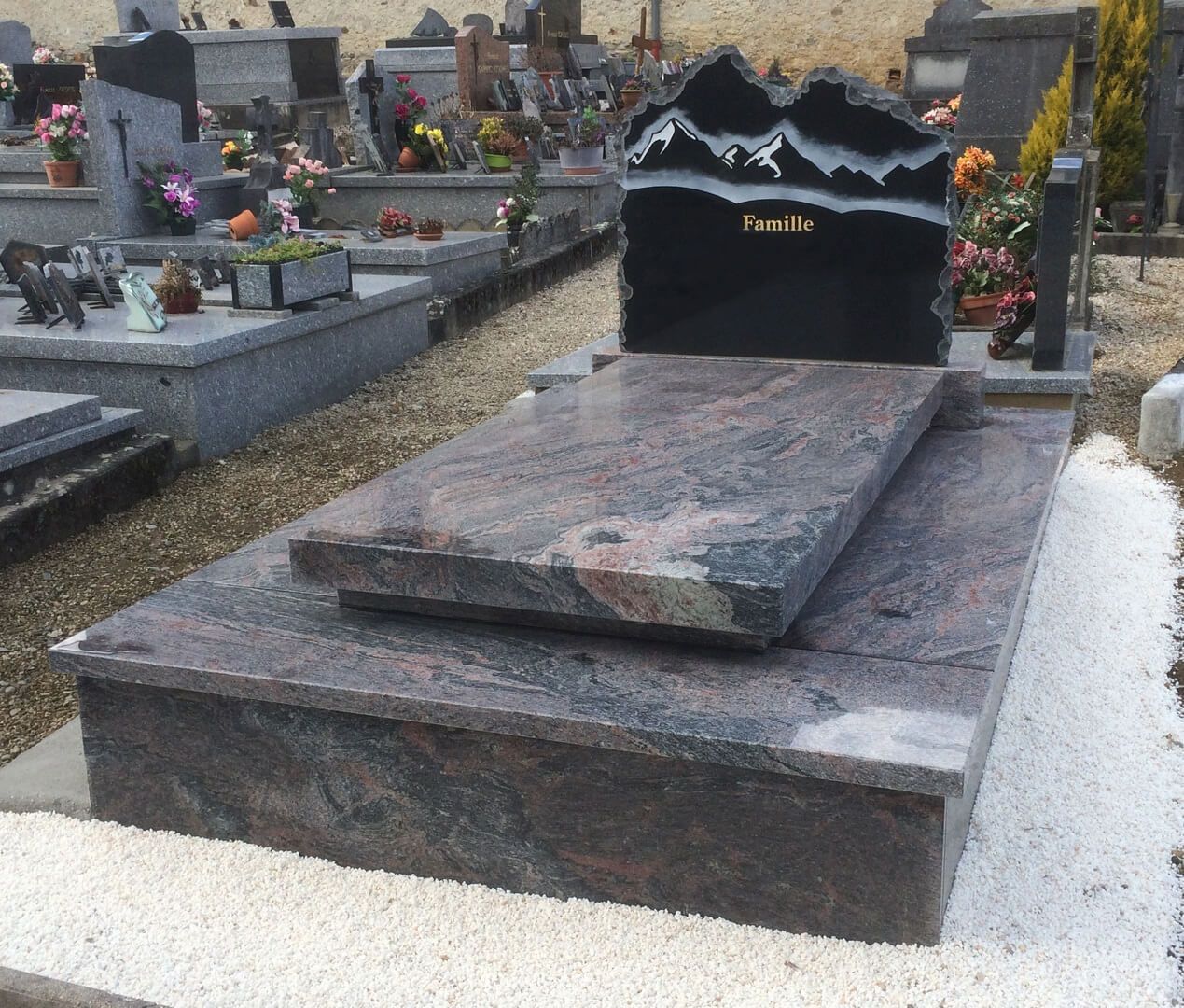 Pierre tombale dans un cimetière, en granit noir et rouge, ornée de motifs de montagne et portant un nom.