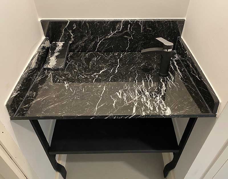 Meuble-lavabo en marbre noir avec lavabo et robinet, soutenu par des pieds et une étagère en métal noir, installé dans une alcôve aux murs blancs.
