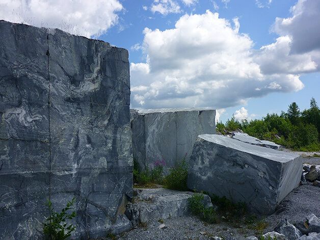 De gros blocs de pierre grise dans une carrière, sous un ciel bleu parsemé de nuages ​​blancs duveteux et de verdure.