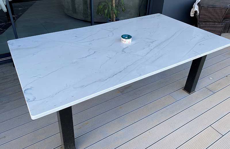 Table d'extérieur rectangulaire avec plateau blanc effet marbre, pieds noirs et un petit objet posé dessus.
