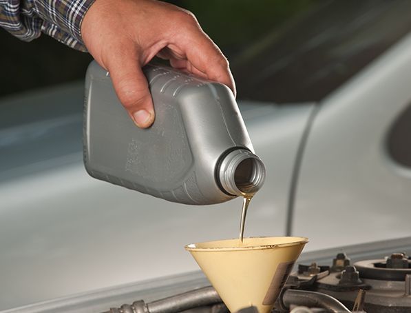 Une main verse de l'huile moteur dorée d'une bouteille en plastique grise dans un entonnoir jaune placé dans le compartiment moteur d'une voiture.