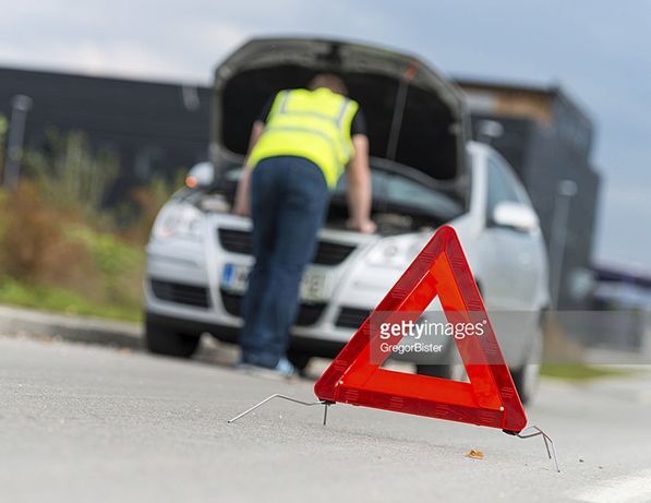 Une personne portant un gilet haute visibilité inspecte le moteur d'une voiture en bord de route, avec un triangle de signalisation rouge au premier plan.