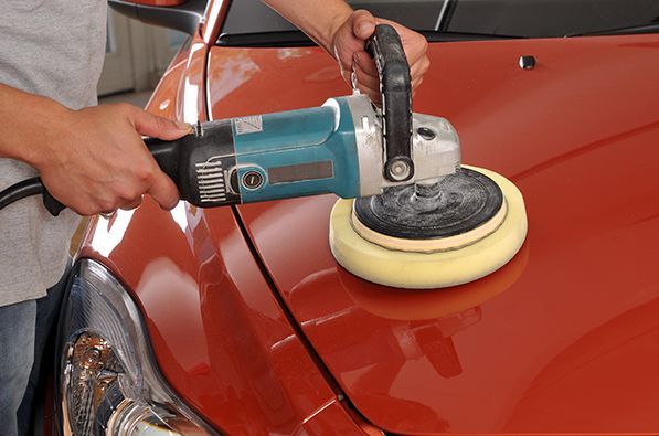 Une personne utilise une polisseuse électrique turquoise avec un tampon en mousse jaune pour polir le capot d'une voiture orange.