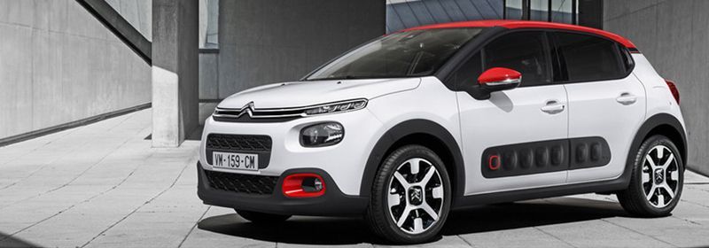 Une Citroën C3 blanche à hayon avec toit rouge, rétroviseurs rouges et protections latérales noires, garée sur du béton.