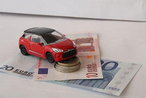 Une petite voiture rouge miniature garée sur une pile de pièces de monnaie posées sur un billet de dix et un billet de vingt euros.