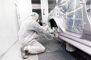 Une personne portant un équipement de protection applique de la peinture en aérosol sur une voiture argentée dans une cabine d'atelier industrielle professionnelle.