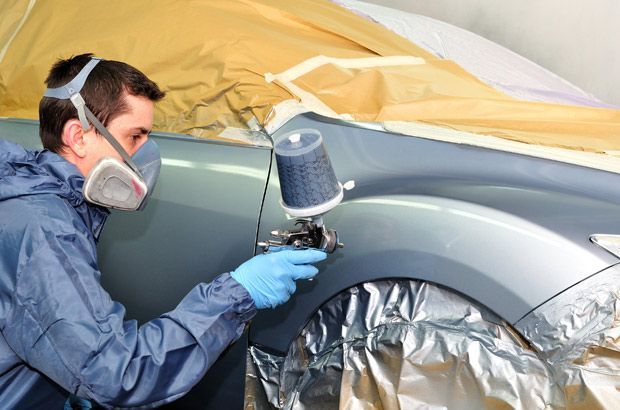 Dans un atelier, une personne portant une combinaison de protection et un masque respiratoire utilise un pistolet à peinture pour peindre l'aile d'une voiture.