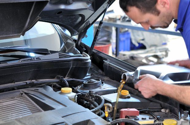 Dans un atelier de réparation, un mécanicien verse de l'huile d'une bouteille en plastique dans le moteur d'une voiture dont le capot est ouvert.