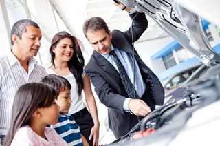 Un vendeur désigne du doigt le moteur d'un véhicule à capot ouvert tout en discutant avec une famille dans une concession automobile.