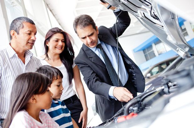 Un vendeur désigne du doigt le moteur d'un véhicule à capot ouvert tout en discutant avec une famille dans une concession automobile.