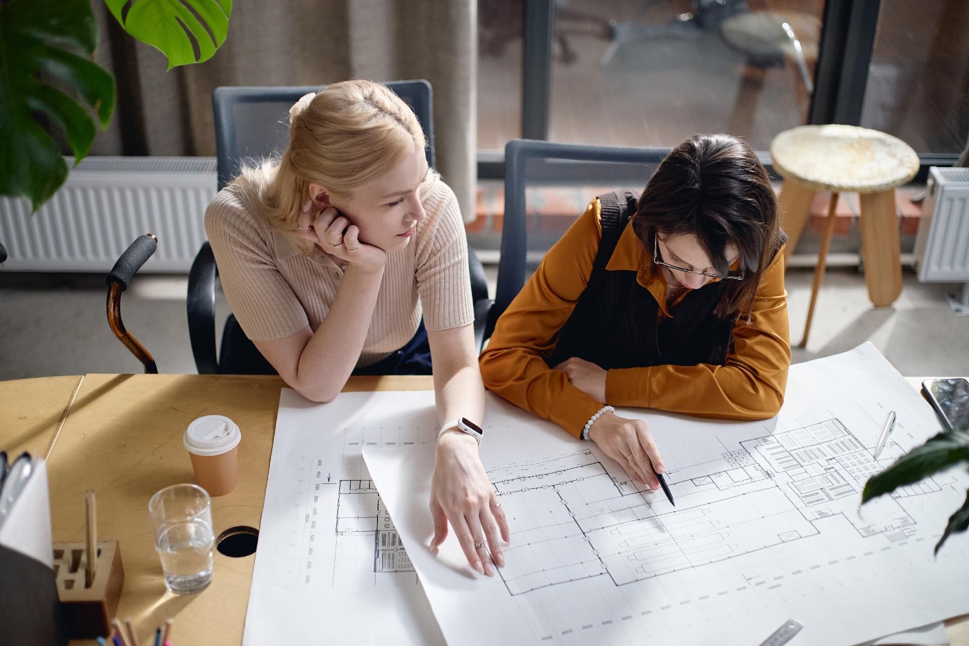 Zwei Personen sitzen an einem Schreibtisch in einem Büro und betrachten Architekturpläne.