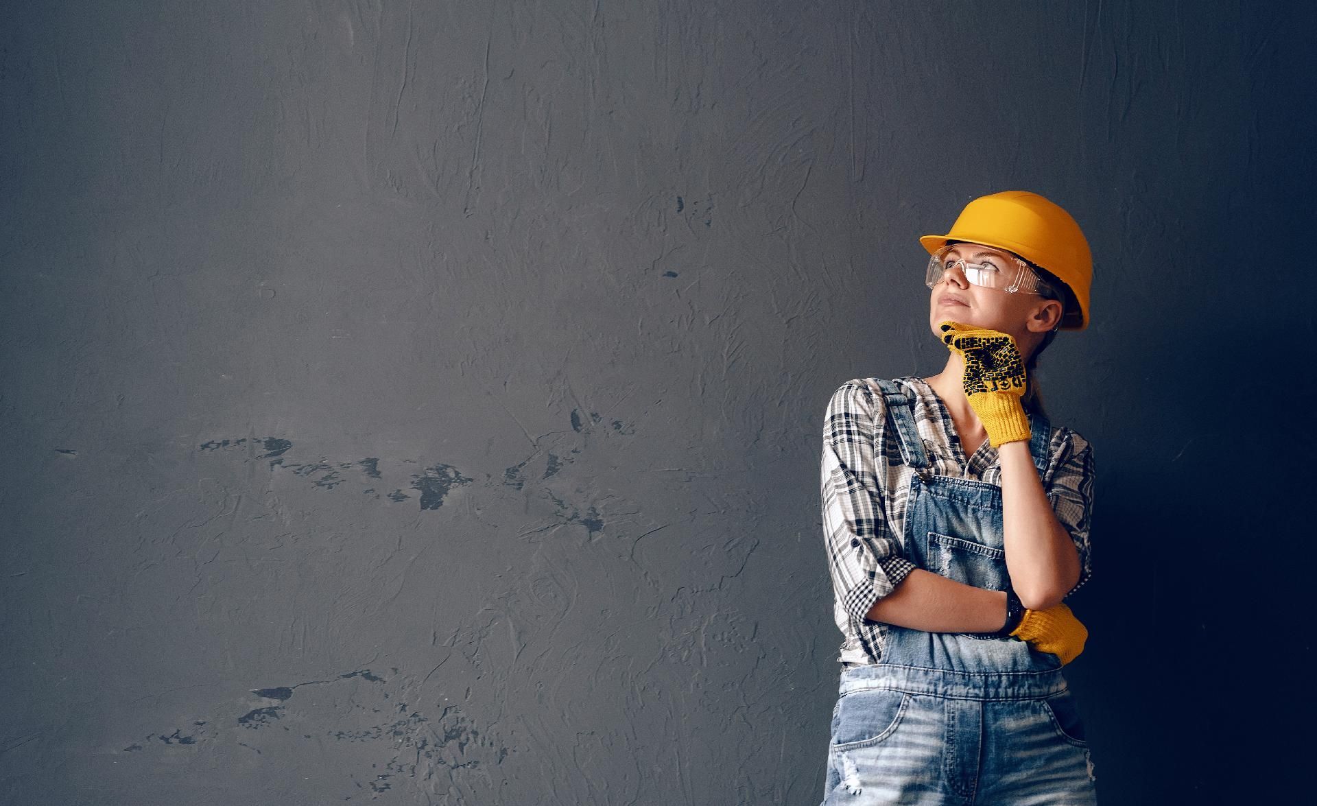 Eine Person mit gelbem Schutzhelm, Schutzbrille und Handschuhen blickt nachdenklich vor einer grauen Wand nach oben.