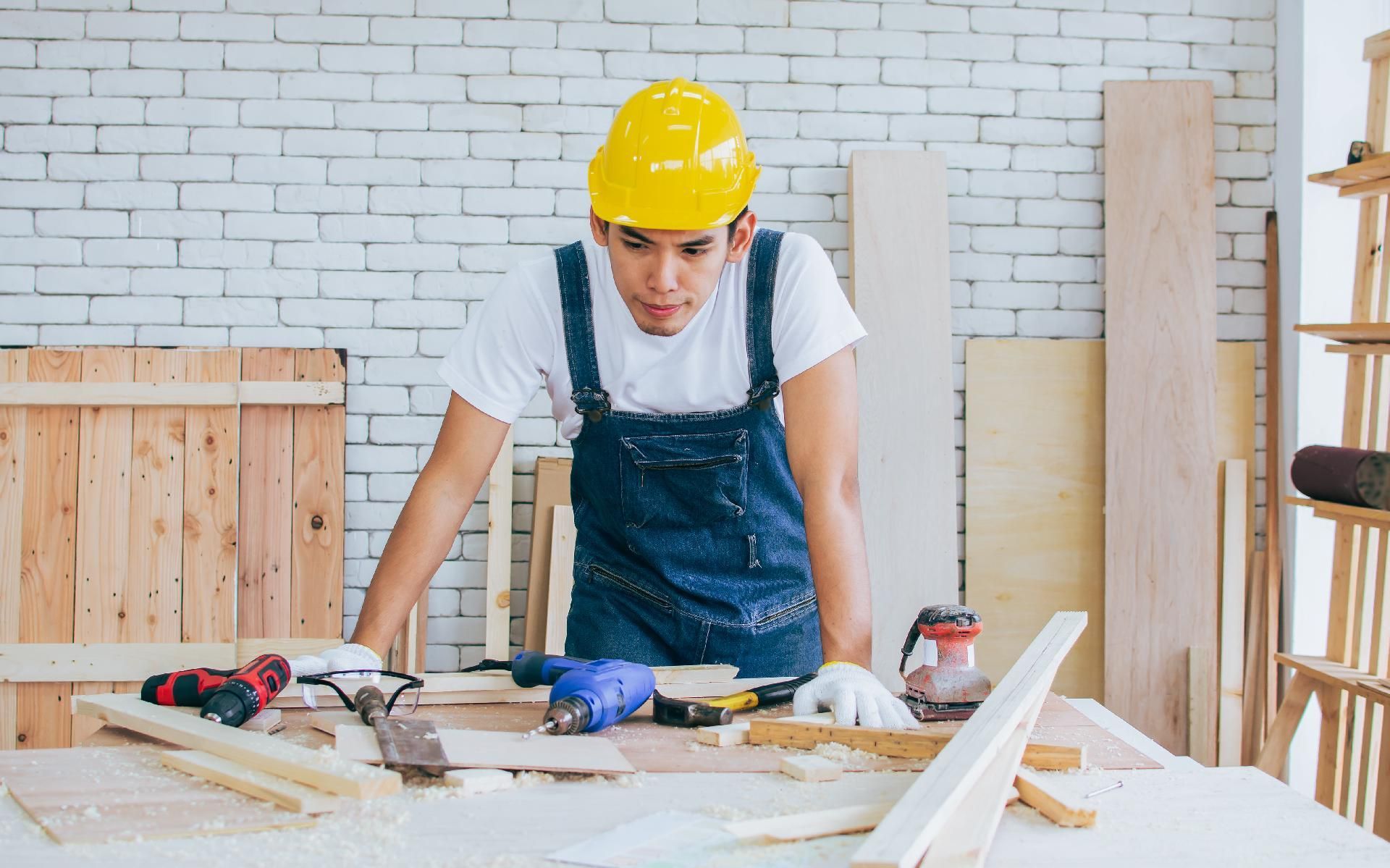 Ein Zimmermann in Schutzhelm und Overall konzentriert sich auf seine Arbeit an einem mit Werkzeug und Holz belegten Tisch.