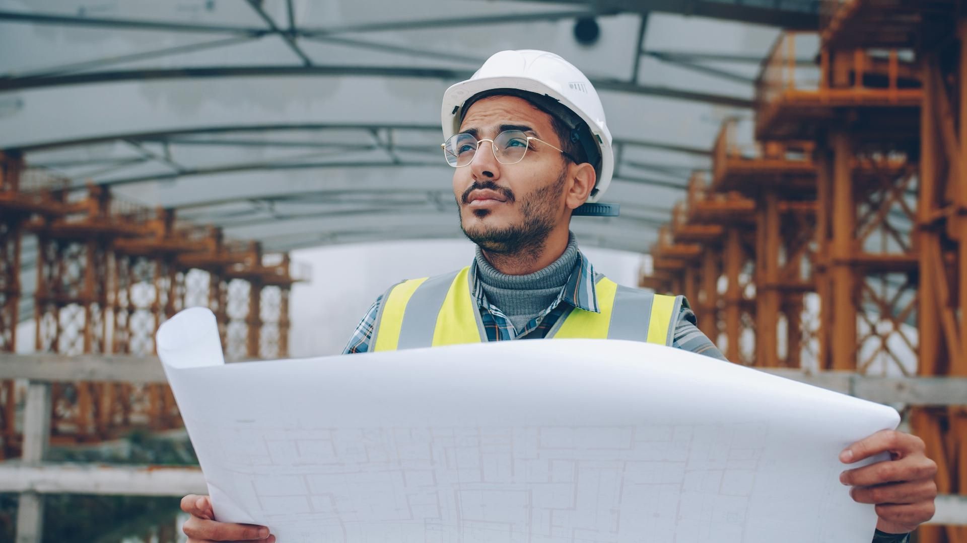Ein Bauarbeiter mit Schutzhelm und Warnweste prüft Baupläne auf einer Baustelle.