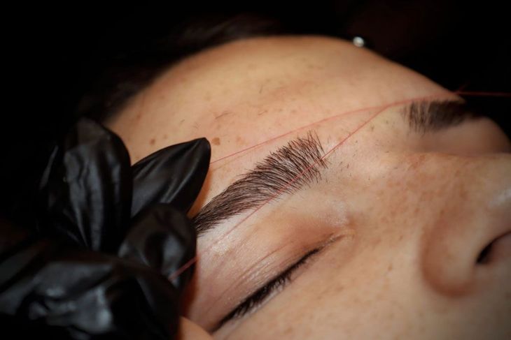 Augenbrauenformen mit Faden, Nahaufnahme. Schwarze behandschuhte Hand, rosa Faden, geschlossene Augen, dunkle Augenbrauen.