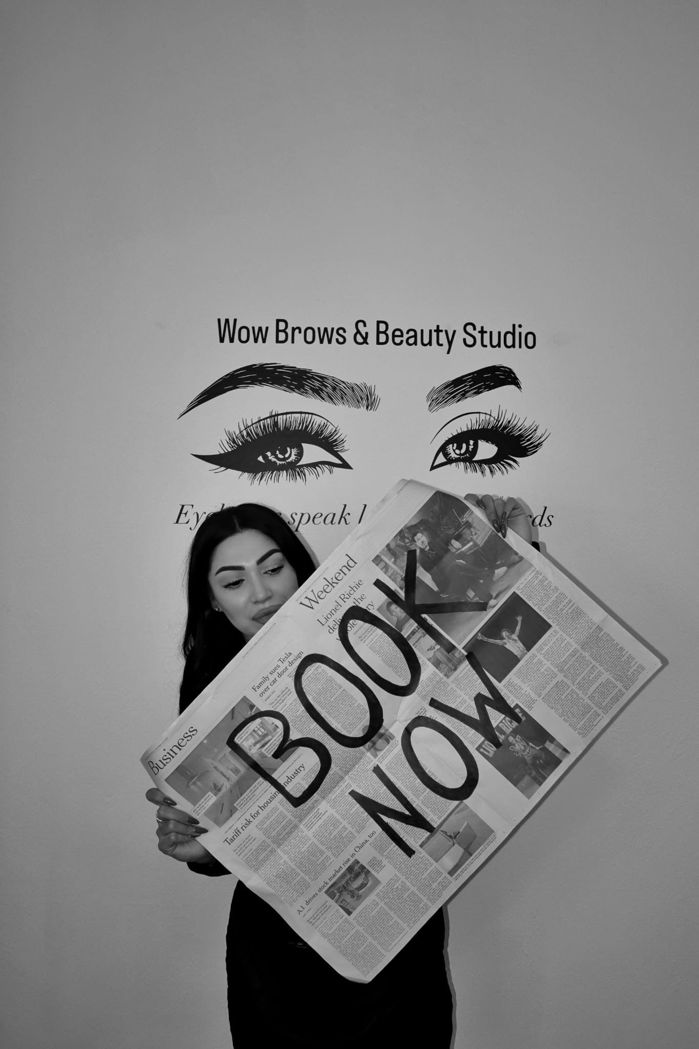 Eine Frau hält eine Zeitung mit der Aufschrift „JETZT BUCHEN“ vor dem Logo eines Schönheitsstudios.