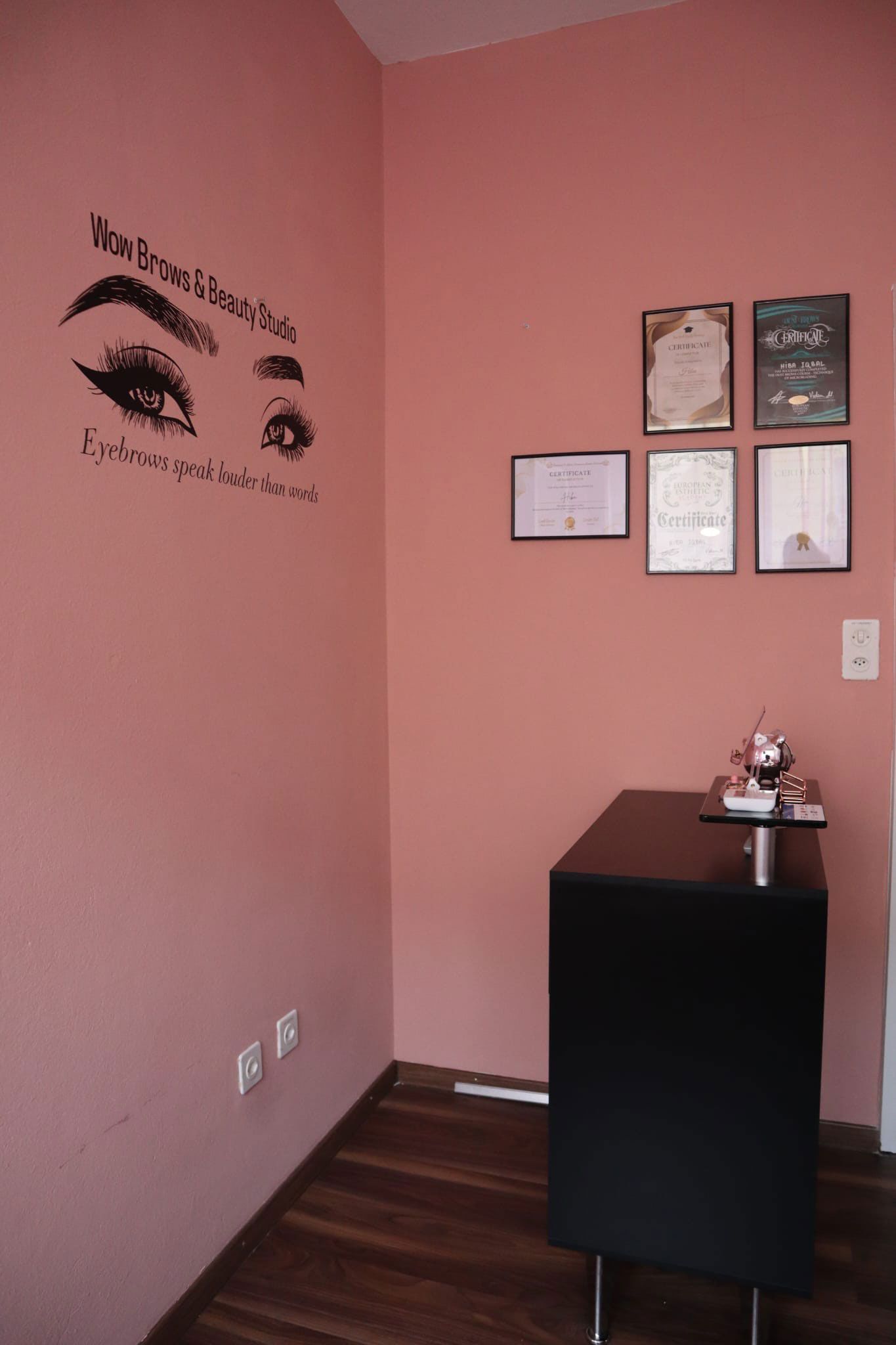 Rosafarbene Ecke mit schwarzem Schrank, Auszeichnungen und einem Augen-Kunstwerk. Holzboden und Dekorationsobjekt auf dem Schrank.