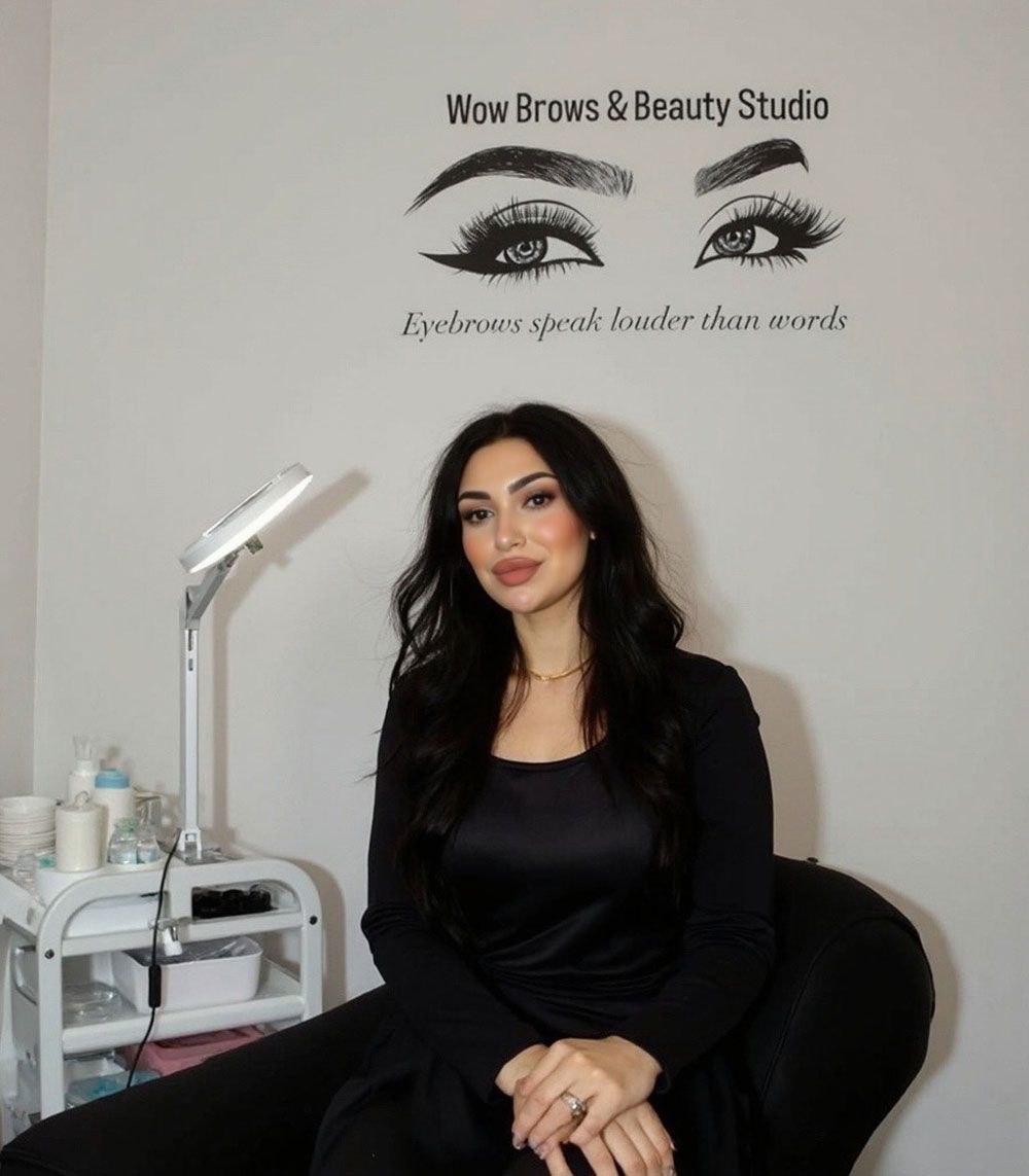 Eine Frau in schwarzem Kleid sitzt in einem Schönheitsstudio, an der Wand hängt das Wow Brows-Logo.