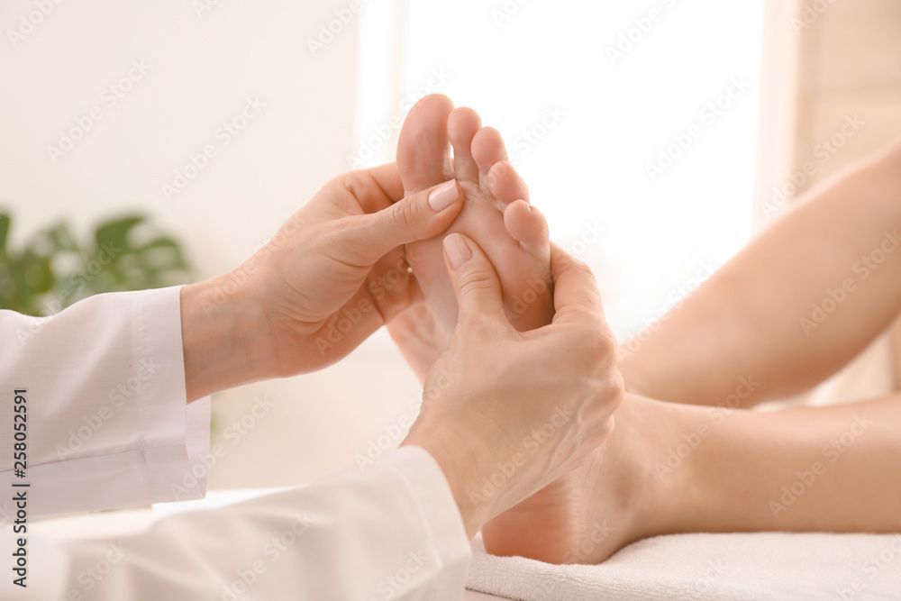 Hände massieren einen Fuss in einem Raum. Bein und Arm der Person sind sichtbar; sie trägt einen weissen Kittel.