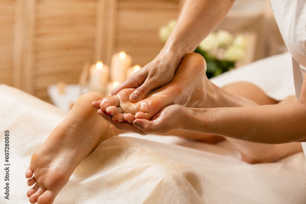 Fussmassage in einem Spa-Ambiente mit Kerzen und Blumen. Hände massieren den eingeölten Fuss.