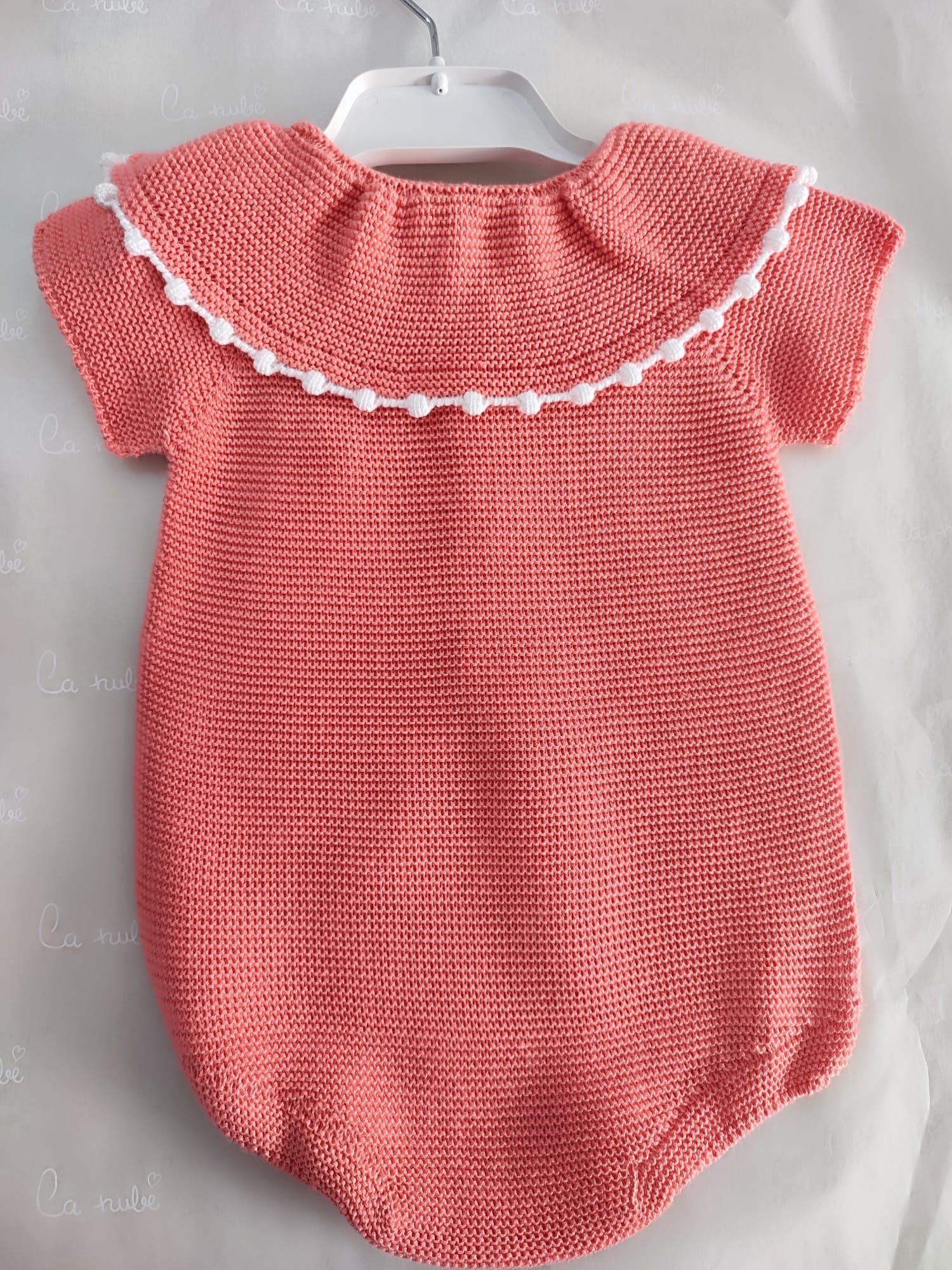 Un suéter rosa con pompones blancos en el cuello está colgado en una percha.