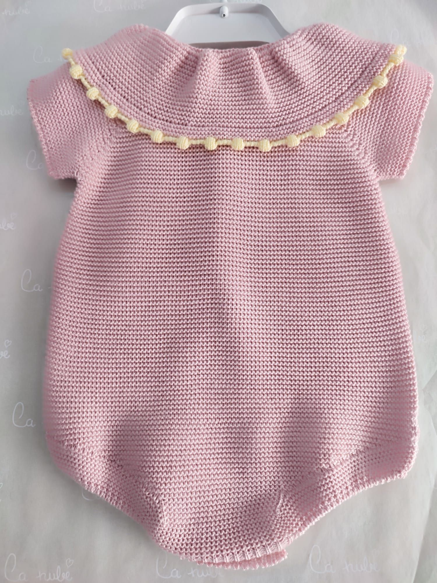 Un mono de bebé de punto rosa está colgado en una percha blanca.