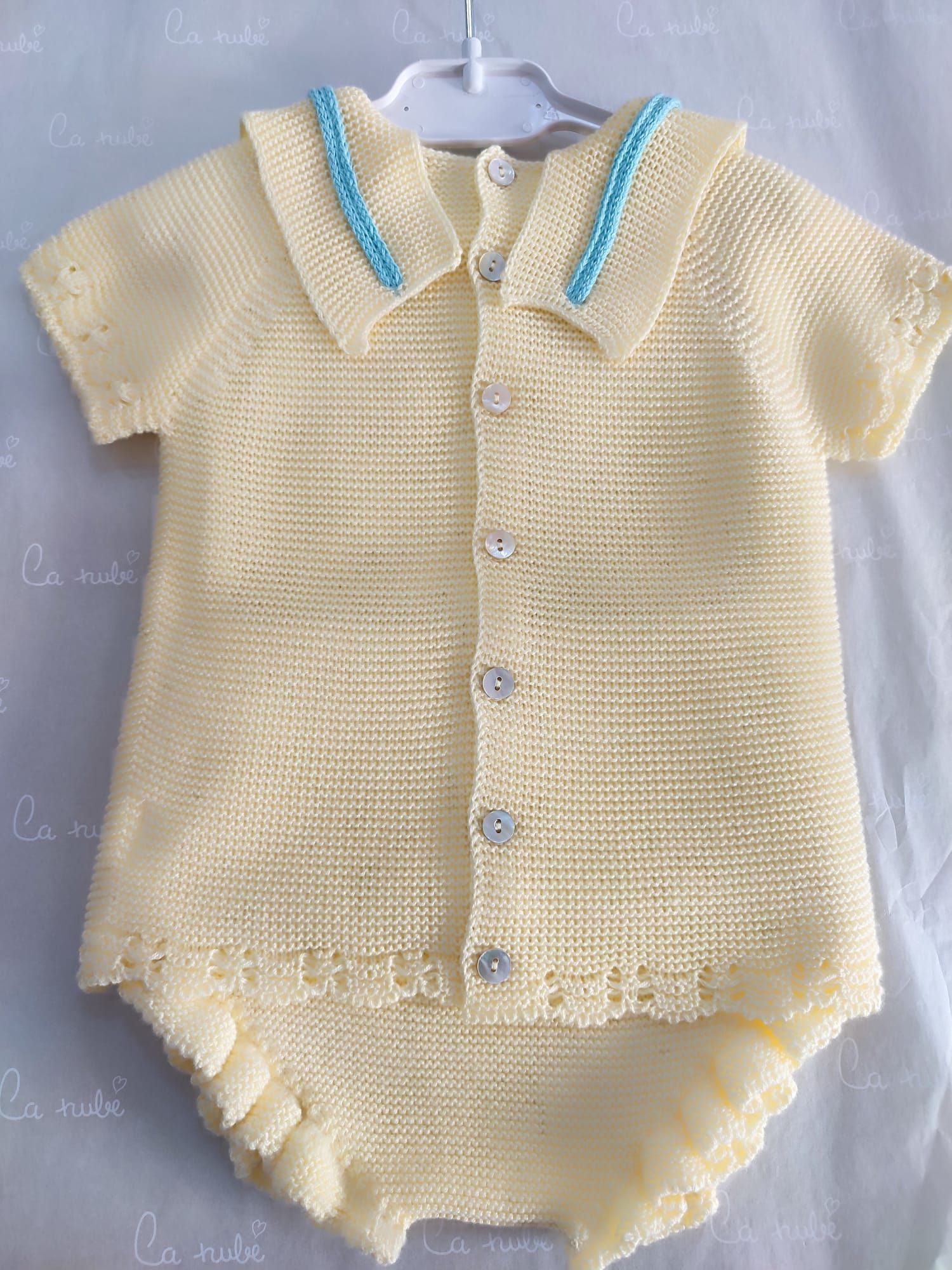 Un conjunto de bebé de punto amarillo con ribete azul está colgado en una percha.