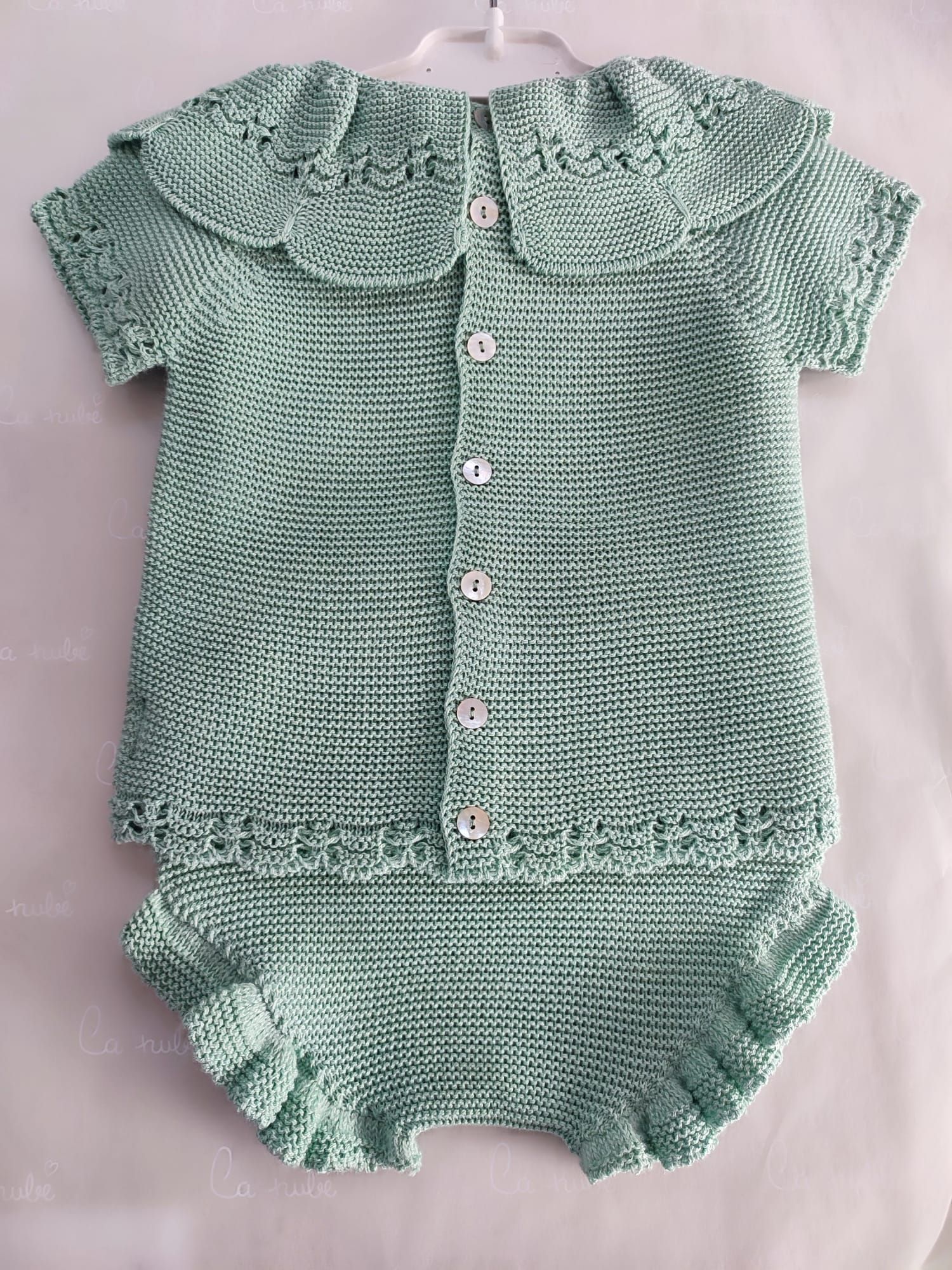 Un conjunto de bebé de punto verde con cuello y botones está colgado en una percha.