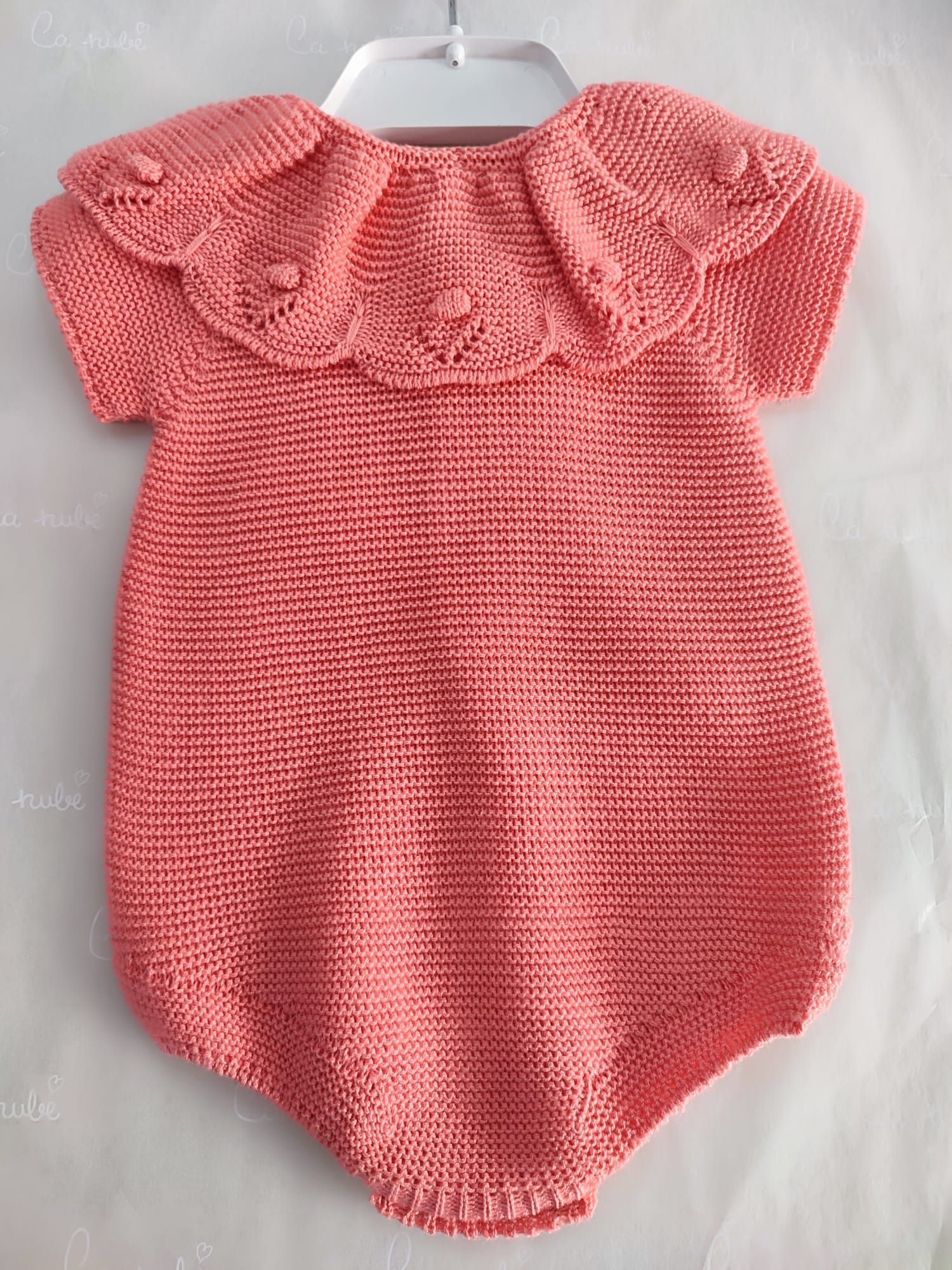 Un mono de bebé de punto rosa con cuello está colgado en una percha blanca.