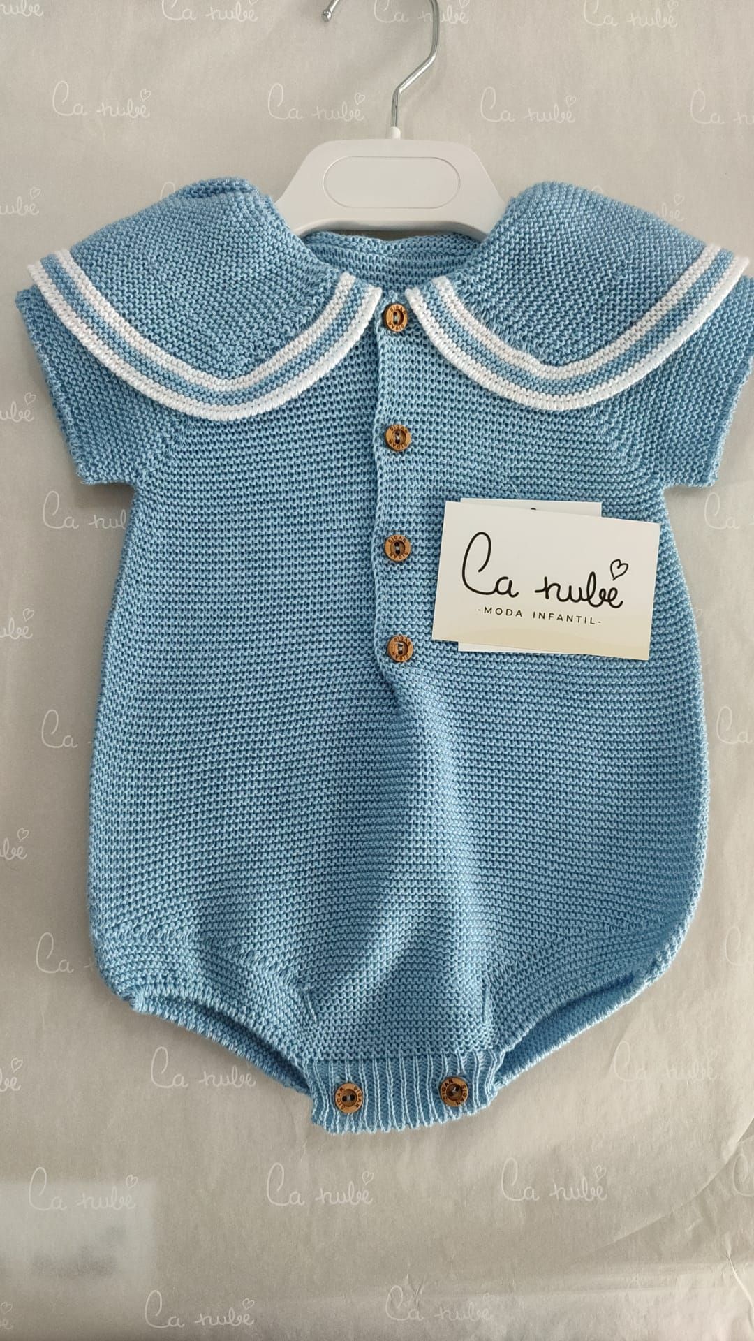 Un mono de bebé de punto azul con cuello y botones blancos está colgado en una percha.