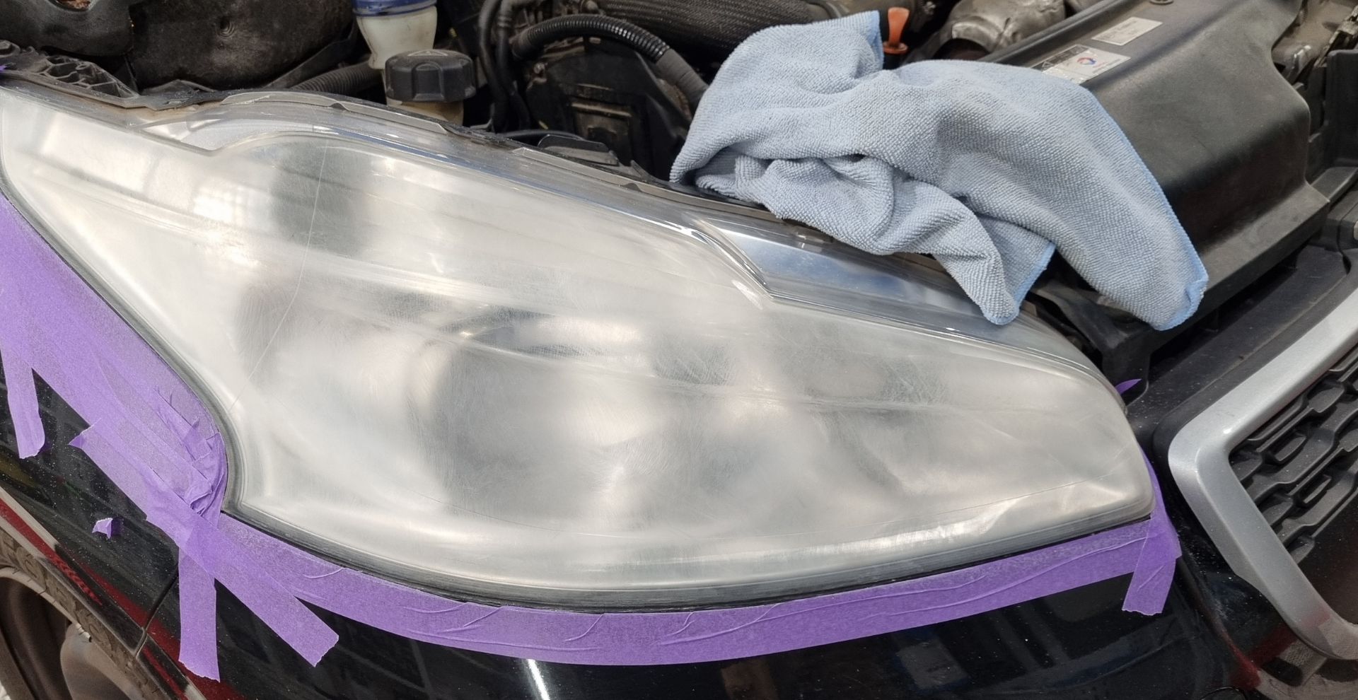 Phare en cours de restauration avec du ruban adhésif, un chiffon et le capot d'une voiture dans un atelier automobile.