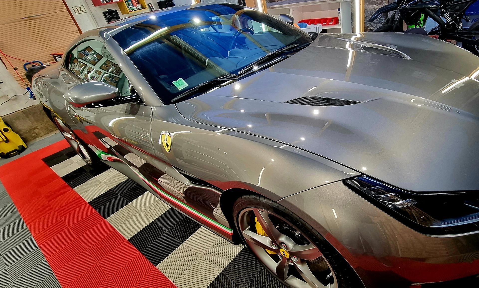 Une voiture de sport Ferrari grise et brillante avec des étriers de frein jaunes garée sur le sol d'un garage à motifs.