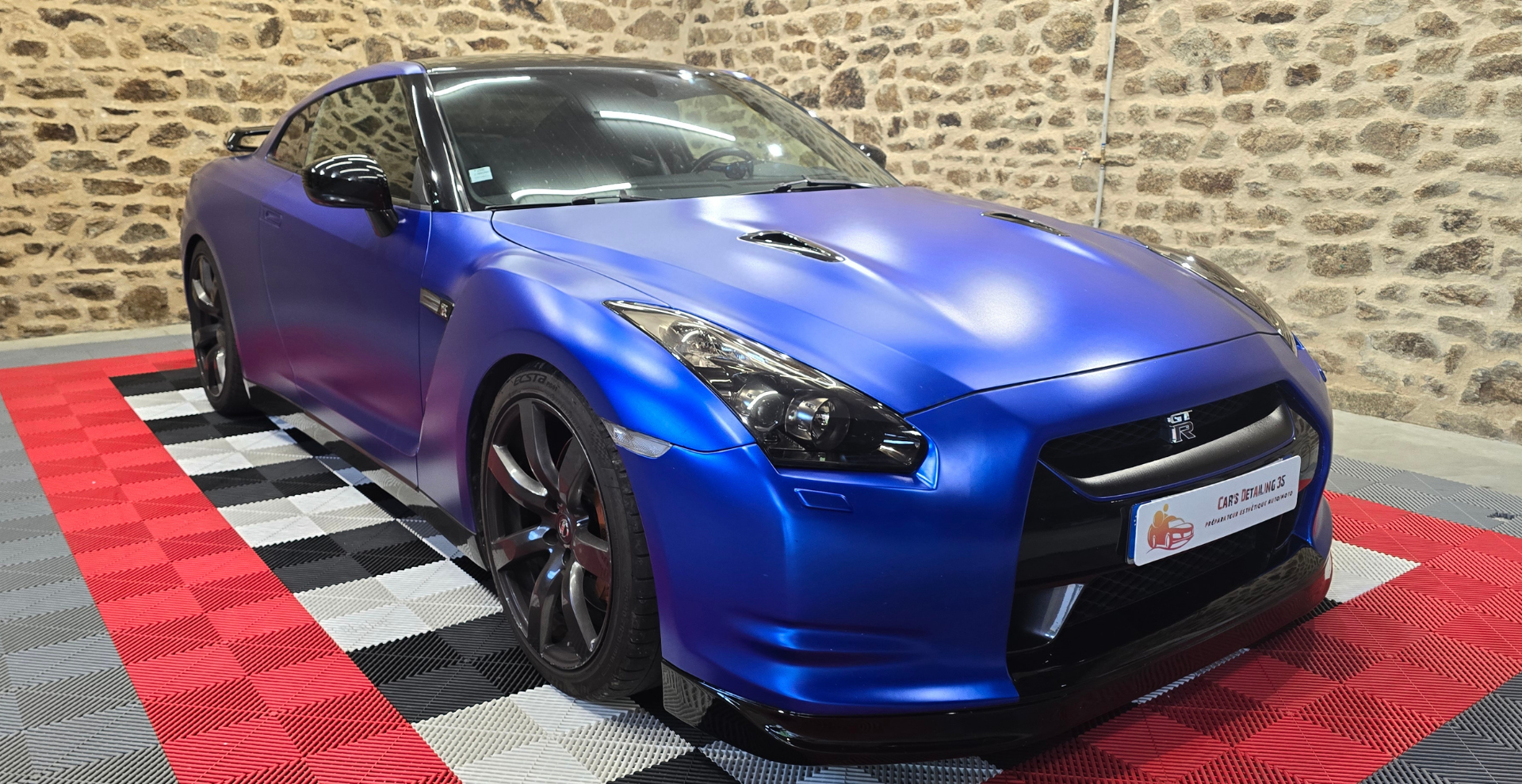 Voiture de sport bleue Nissan GT-R garée sur un sol en damier contre un mur de briques.