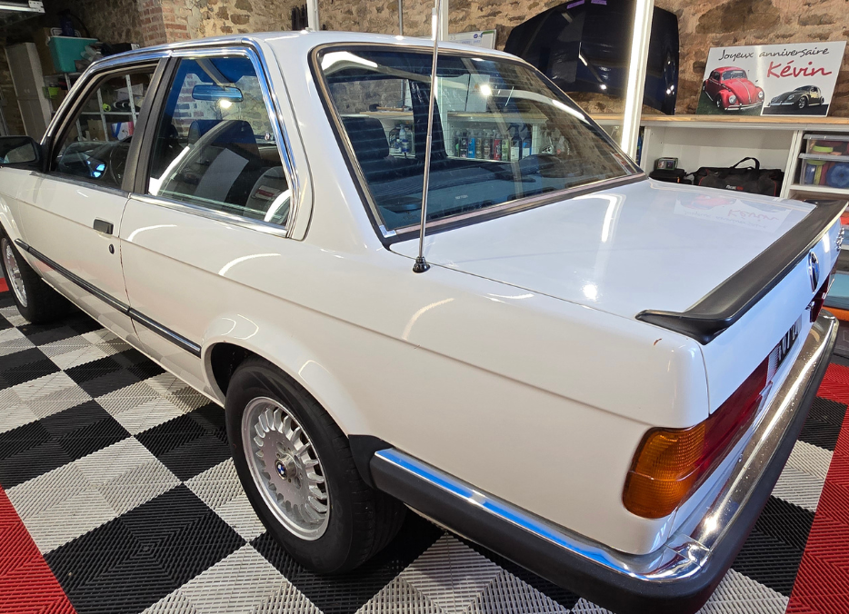 Coupé BMW classique blanc garé dans un garage avec plancher à carreaux, doté d'un becquet noir et de garnitures chromées.